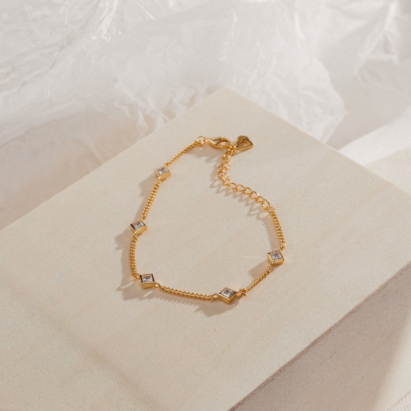 The Venus Diamond Station Bracelet features a gold chain with delicate diamond-shaped accents, ideal as wedding jewelry, shown on a beige textured background.
