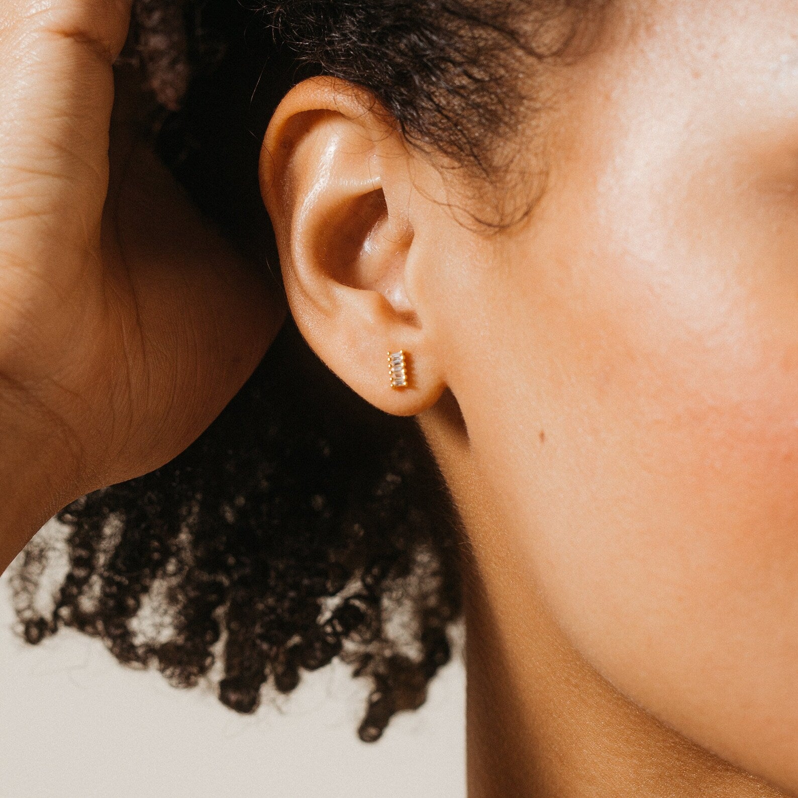 A close-up of a woman's ear wearing Serena Baguette Studs—minimalist stud earrings with small rectangular gold and baguette diamond accents.