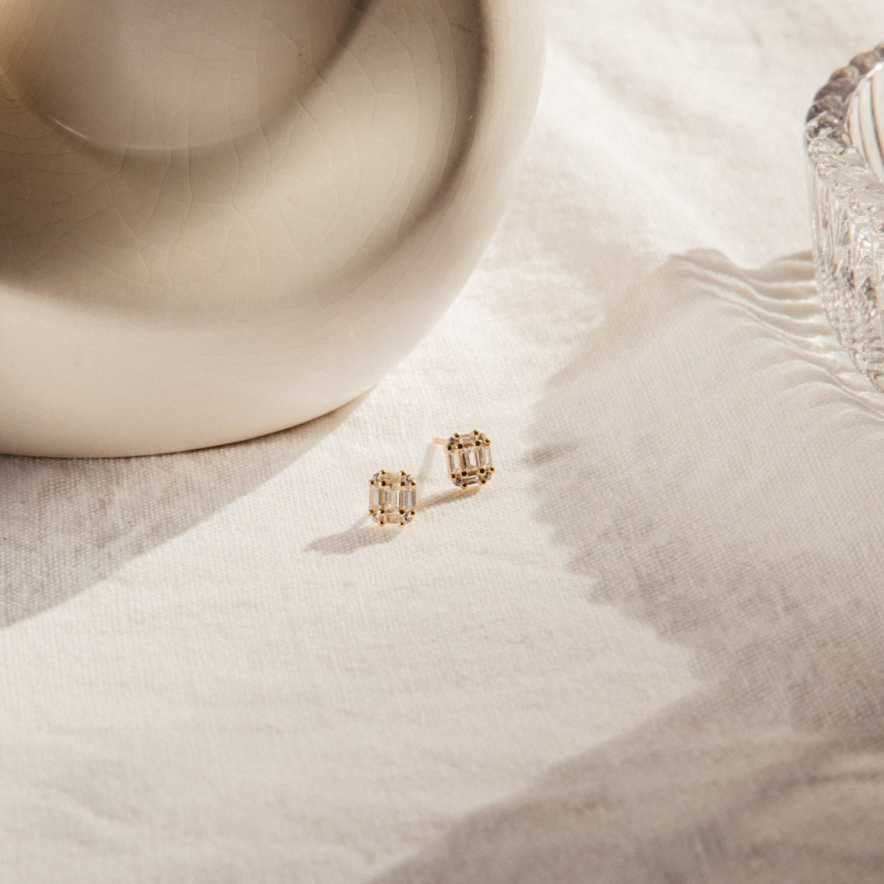The Arabella Diamond Studs, featuring clear CZ stones in an Art Deco design, rest on white textured fabric beside a ceramic object and a glass dish, gently illuminated by natural light.