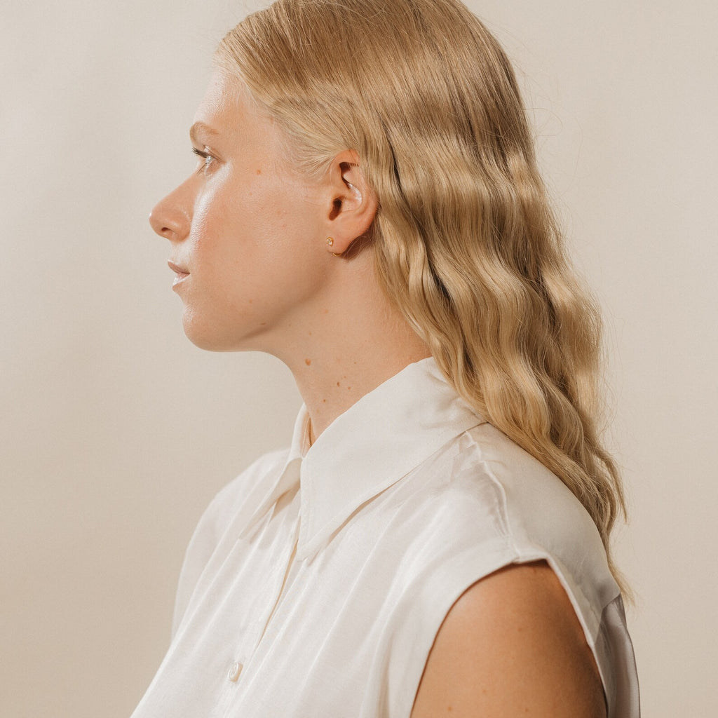 A woman with wavy blonde hair and a white sleeveless blouse, shown in profile against a plain background, wears Saylor III Huggies for an effortlessly chic, everyday look.