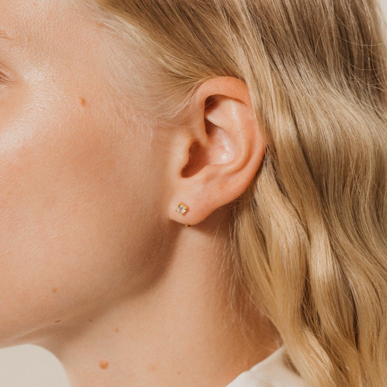 Close-up of a woman's ear wearing Saylor III Huggies, stylish small gold earrings ideal for everyday wear, with wavy blonde hair.