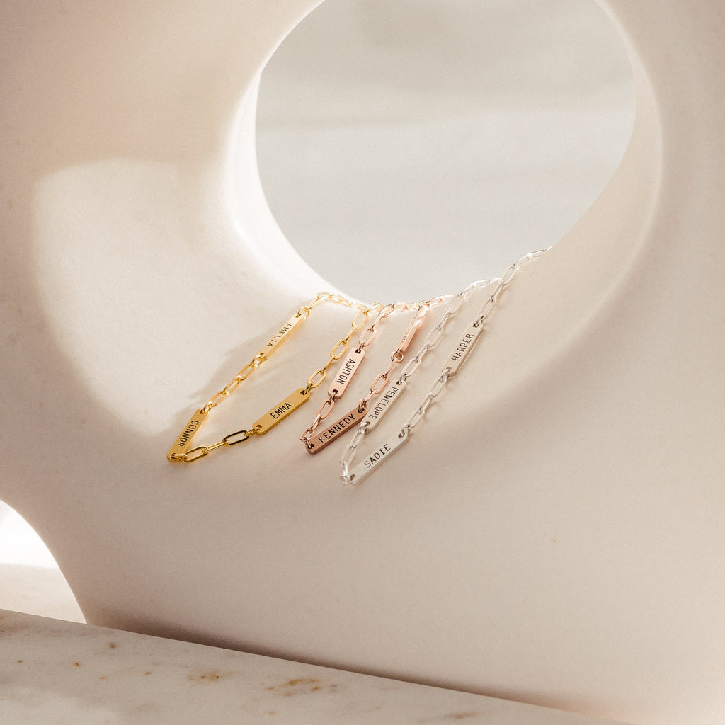 Four delicate chain bracelets, including the Mini Name Bar Bracelet and a stylish paperclip style, in gold, rose gold, and silver are displayed on a curved white sculpture.