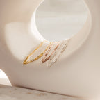 Four delicate chain bracelets, including the Mini Name Bar Bracelet and a stylish paperclip style, in gold, rose gold, and silver are displayed on a curved white sculpture.