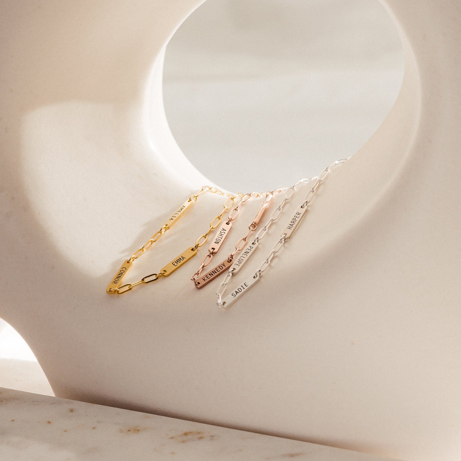 Four delicate chain bracelets, including the Mini Name Bar Bracelet and a stylish paperclip style, in gold, rose gold, and silver are displayed on a curved white sculpture.