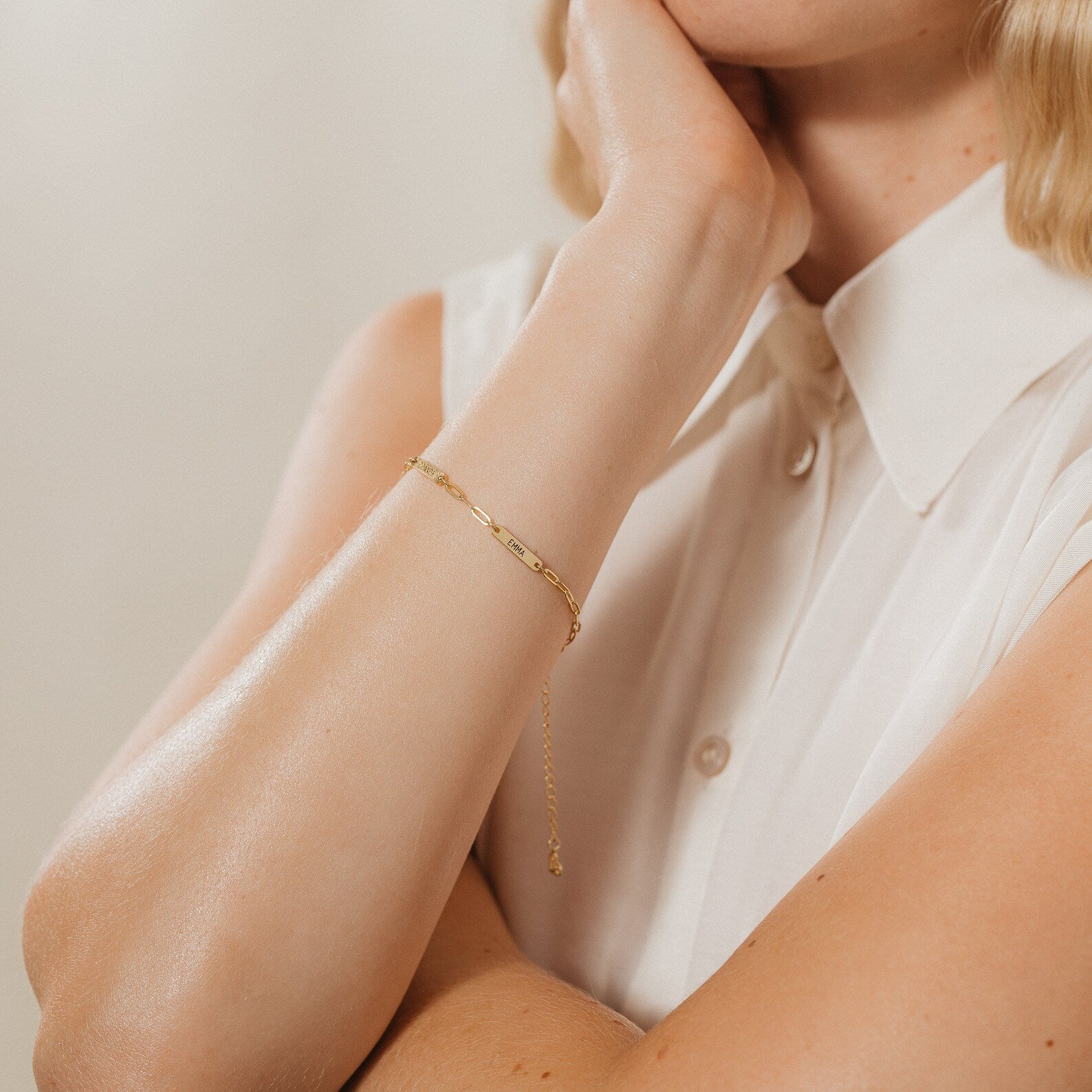 A woman in a sleeveless white blouse rests her chin on her hand, wearing the Mini Name Bar Bracelet.