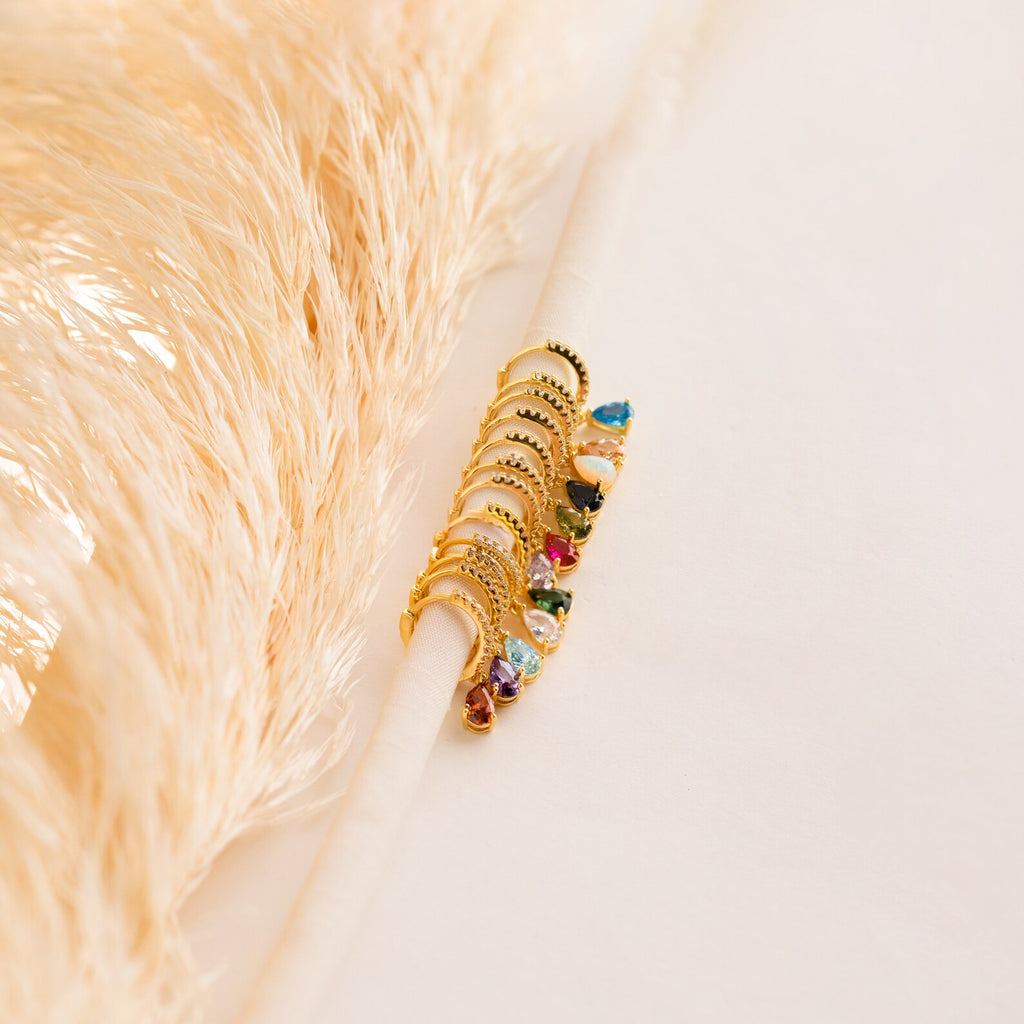 Pave Birthstone Drop Huggies with colorful gemstones hang on a white rod, pampas grass in the background, evoking elegance and the chic style of gemstone hoop earrings.