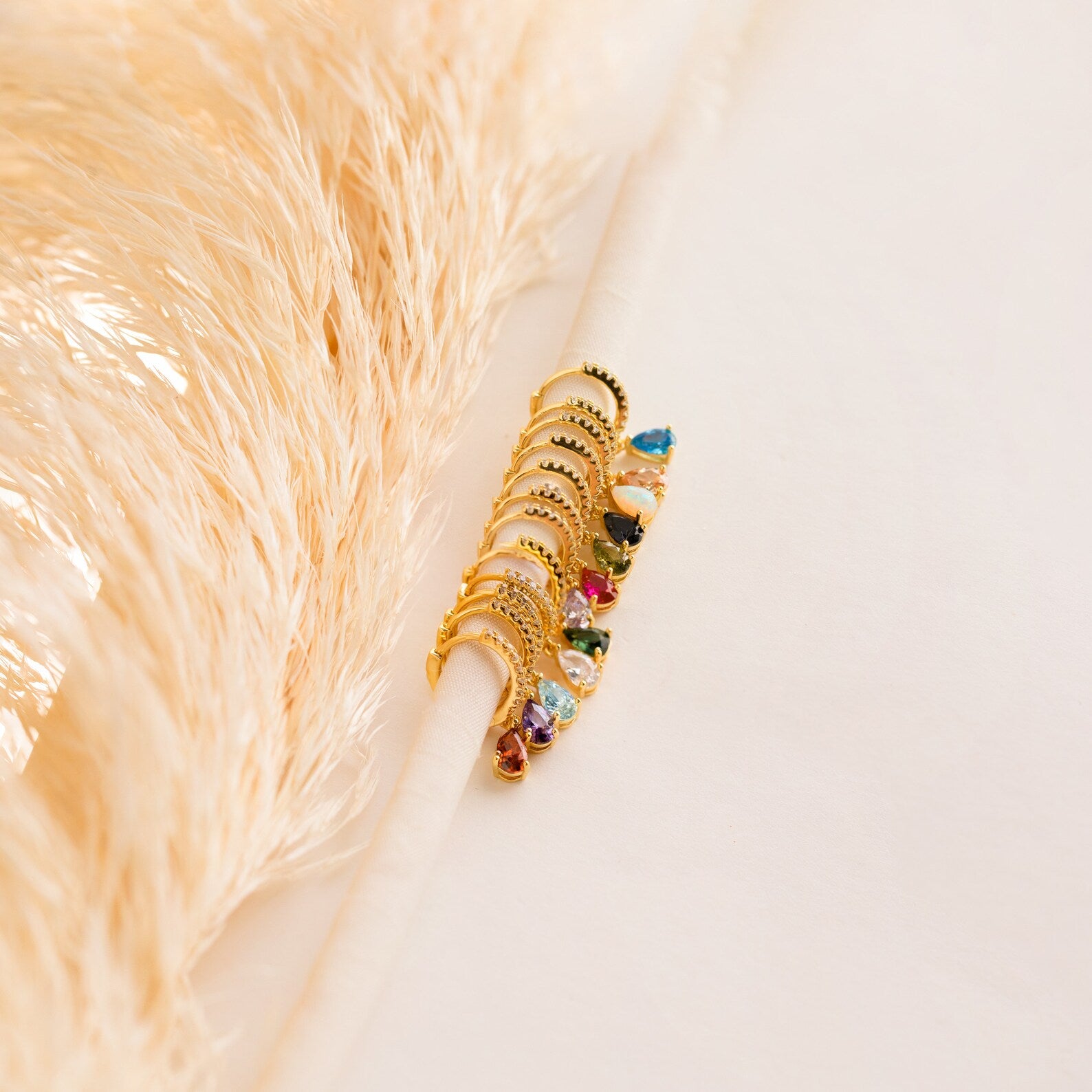 Pave Birthstone Drop Huggies with colorful gemstones hang on a white rod, pampas grass in the background, evoking elegance and the chic style of gemstone hoop earrings.