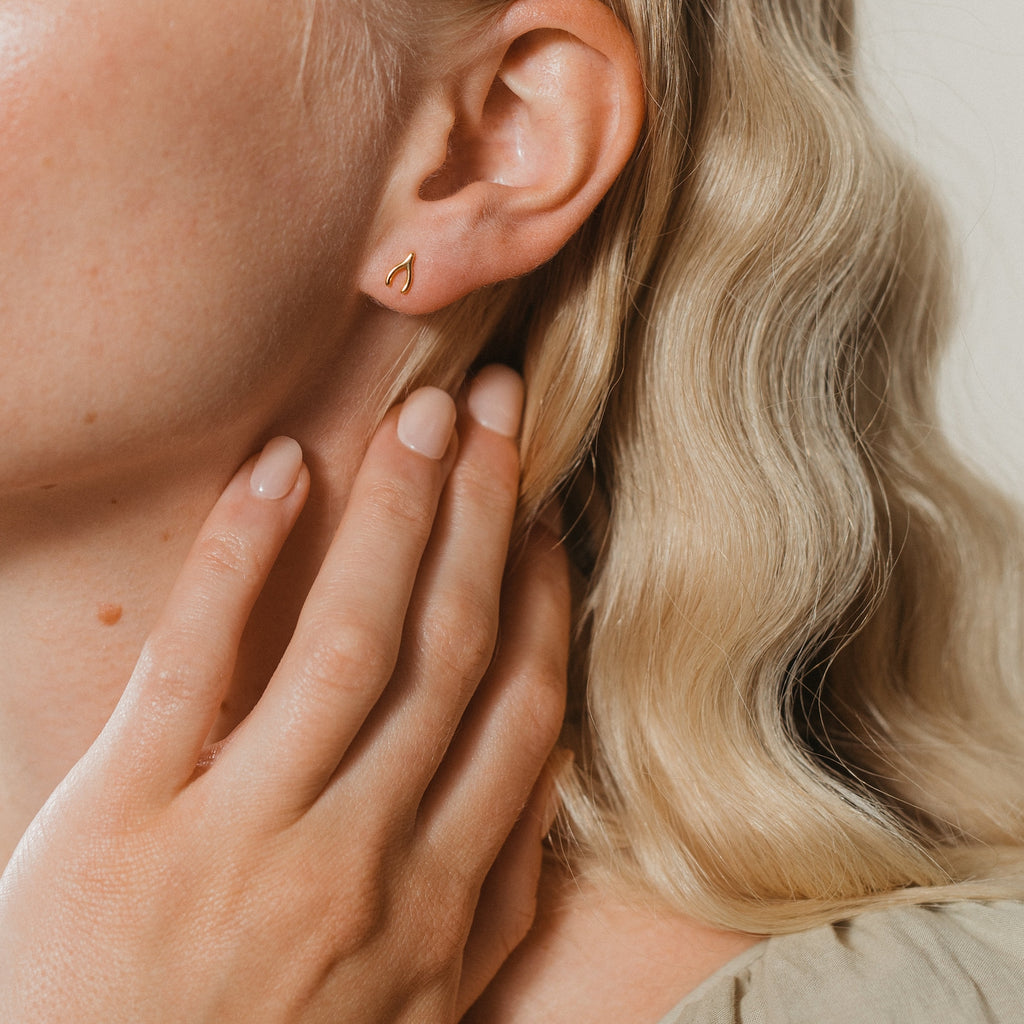 A woman with wavy blonde hair wears a small gold earring from the Lucky Studs Set and touches her neck with her hand.