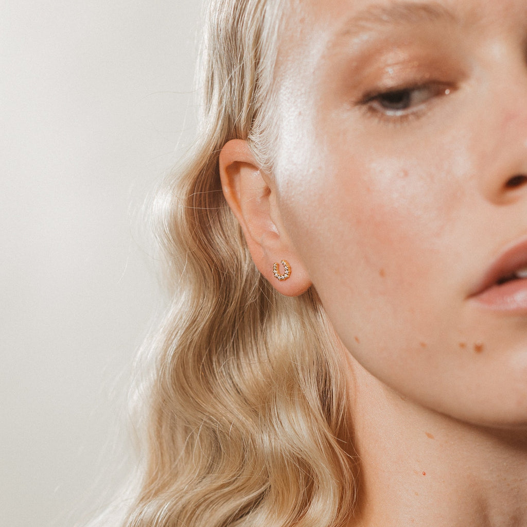 Close-up of a woman with wavy blonde hair wearing the Lucky Studs Set: a small gold hoop and a delicate wishbone earring in her left ear.