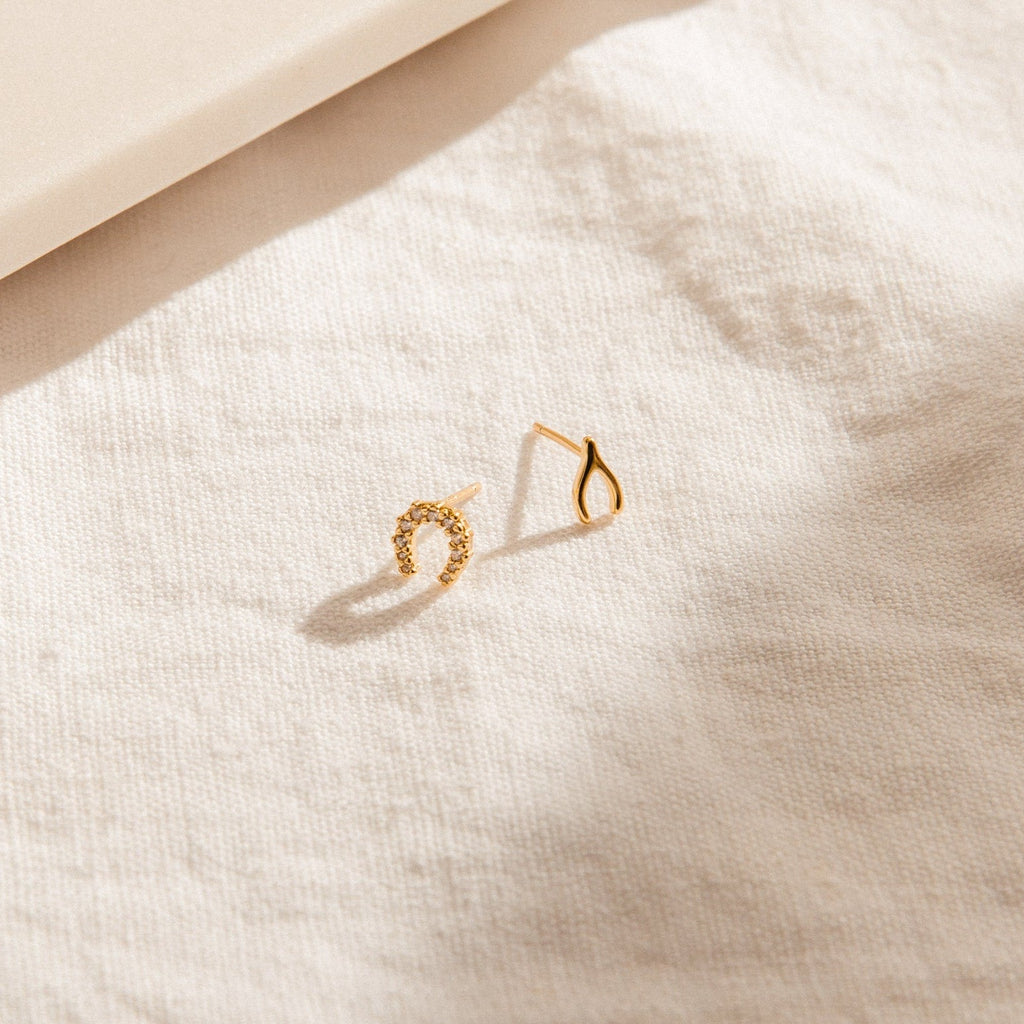 The Lucky Studs Set features two delicate mismatched stud earrings—one is a sparkling diamond horseshoe, and the other a graceful gold teardrop, resting on textured white fabric.