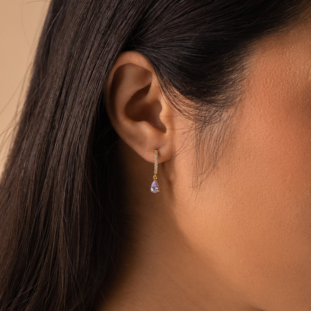 Close-up of a woman's ear wearing Pave Birthstone Drop Huggies, featuring delicate gold pavé diamond hoops with a small purple gemstone drop.
