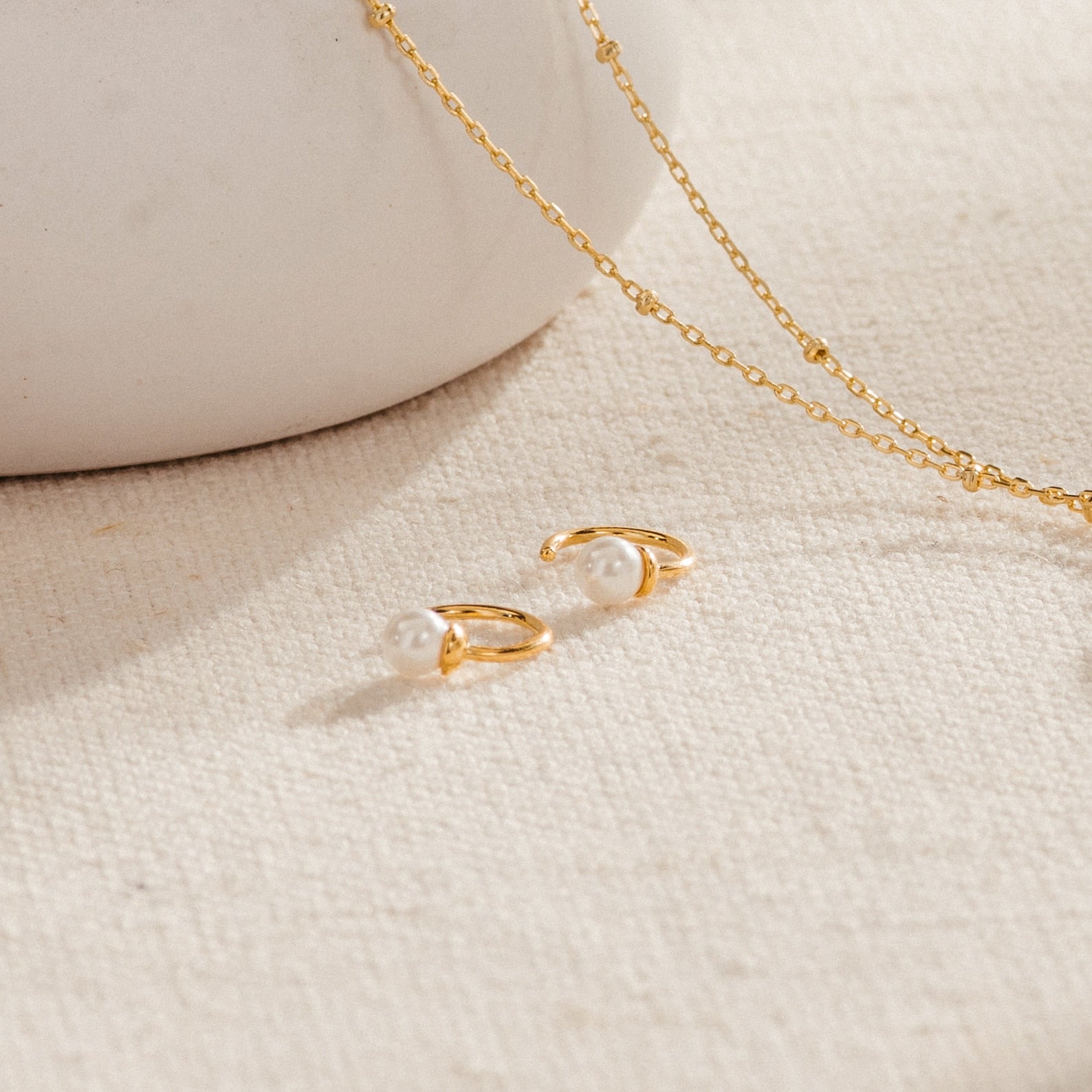 Lila Pearl Huggies and dainty hoop earrings with pearls, plus a gold chain necklace, are styled on a textured beige surface for a modern jewelry vibe.