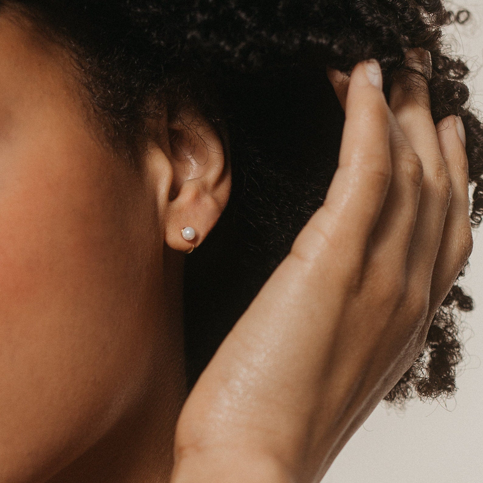 Close-up of a woman touching her curly hair, wearing Lila Pearl Huggies that add a touch of modern jewelry charm to her look.