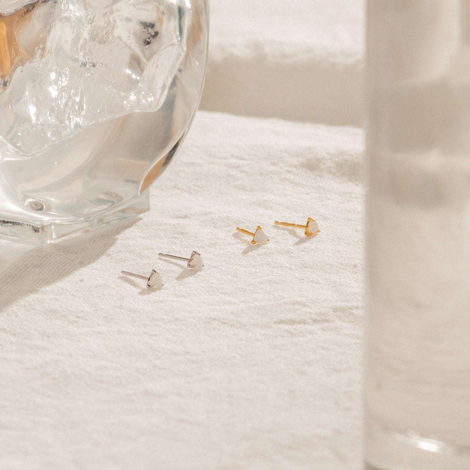 Two pairs of Opal Heart Studs rest on a white cloth beside a glass of ice water—perfect for adding minimalistic glam to your everyday look.