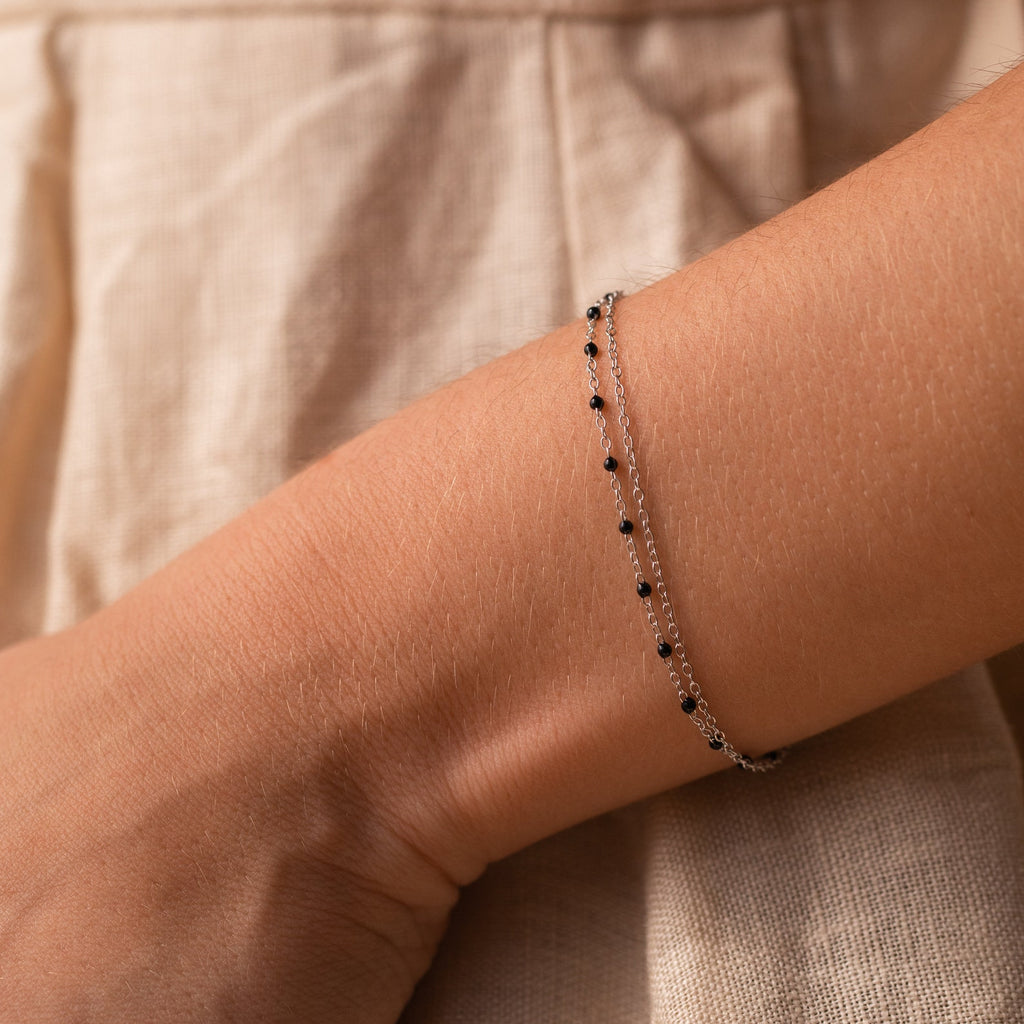 The Calypso Station Bracelet, a delicate silver boho piece with small black beads, rests on a wrist—ideal for stacking accessories—set against a beige fabric background.