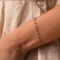 The Calypso Station Bracelet, a delicate silver boho piece with small black beads, rests on a wrist—ideal for stacking accessories—set against a beige fabric background.