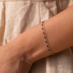 The Calypso Station Bracelet, a delicate silver boho piece with small black beads, rests on a wrist—ideal for stacking accessories—set against a beige fabric background.