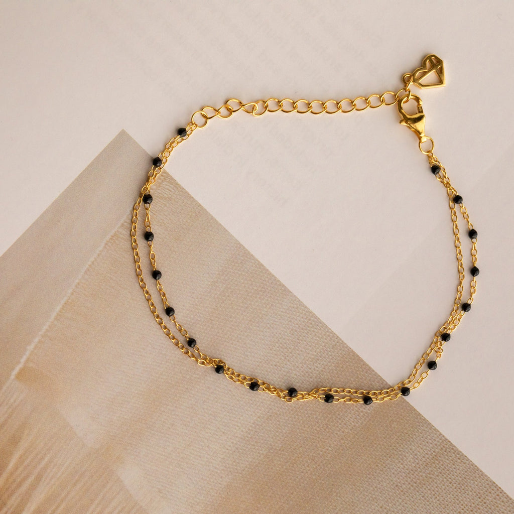 The Calypso Station Bracelet is a delicate gold bracelet with small black beads and a heart-shaped charm, ideal for stacking or pairing with your favorite boho accessories, shown on a neutral background.
