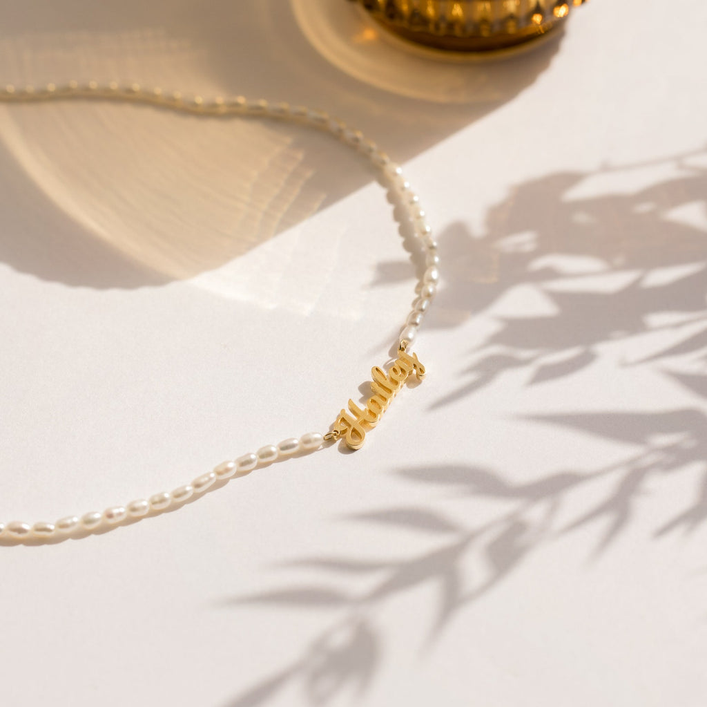 The Mellow Pearl Name Necklace, featuring a dainty nameplate charm and gold "mama" pendant, is displayed on a white surface with gentle shadows.