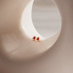 Ruby Heart Studs, two small ruby heart earrings, are displayed on a smooth curved beige surface with a round opening—perfect date night accessories to add romantic charm to your look.