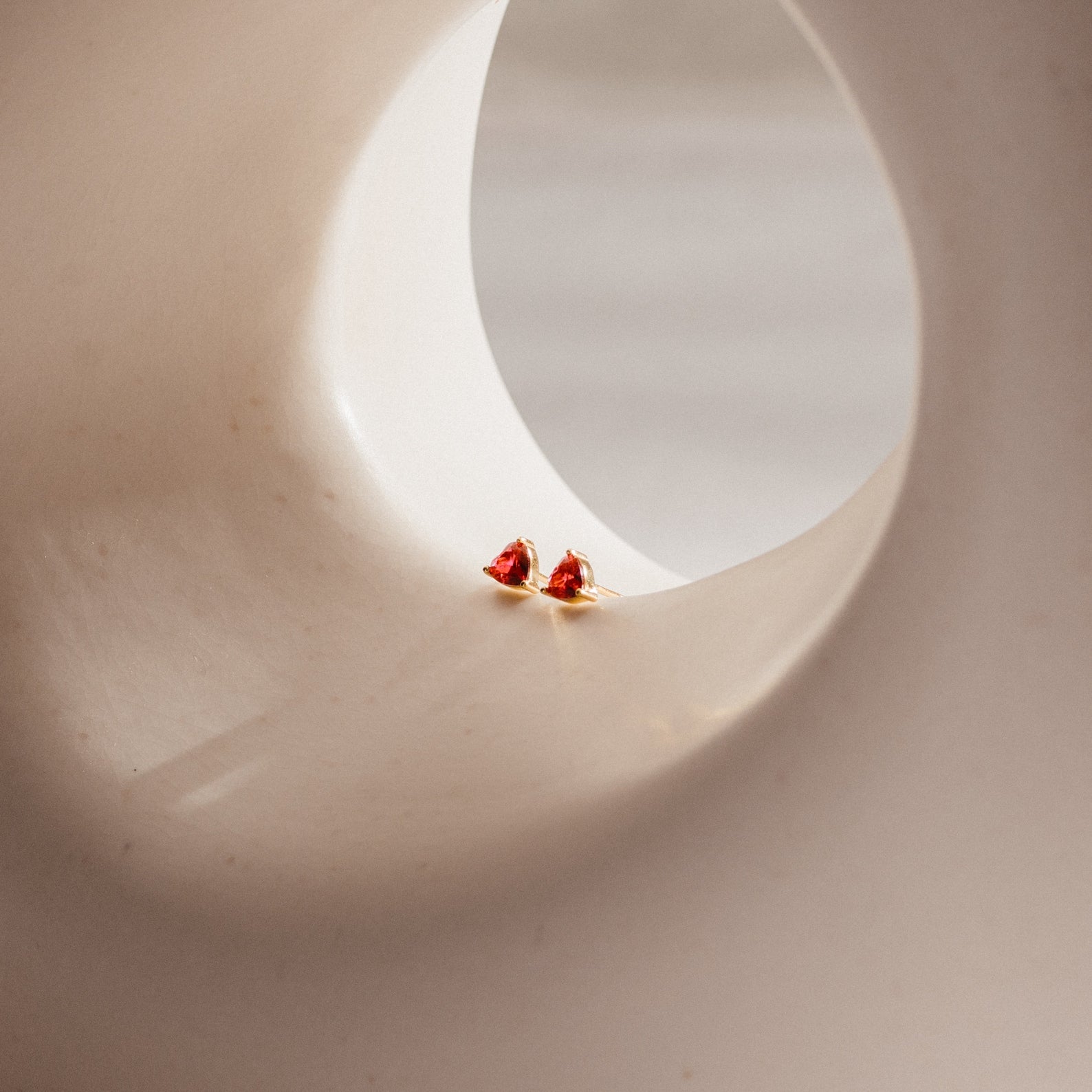 Ruby Heart Studs, two small ruby heart earrings, are displayed on a smooth curved beige surface with a round opening—perfect date night accessories to add romantic charm to your look.