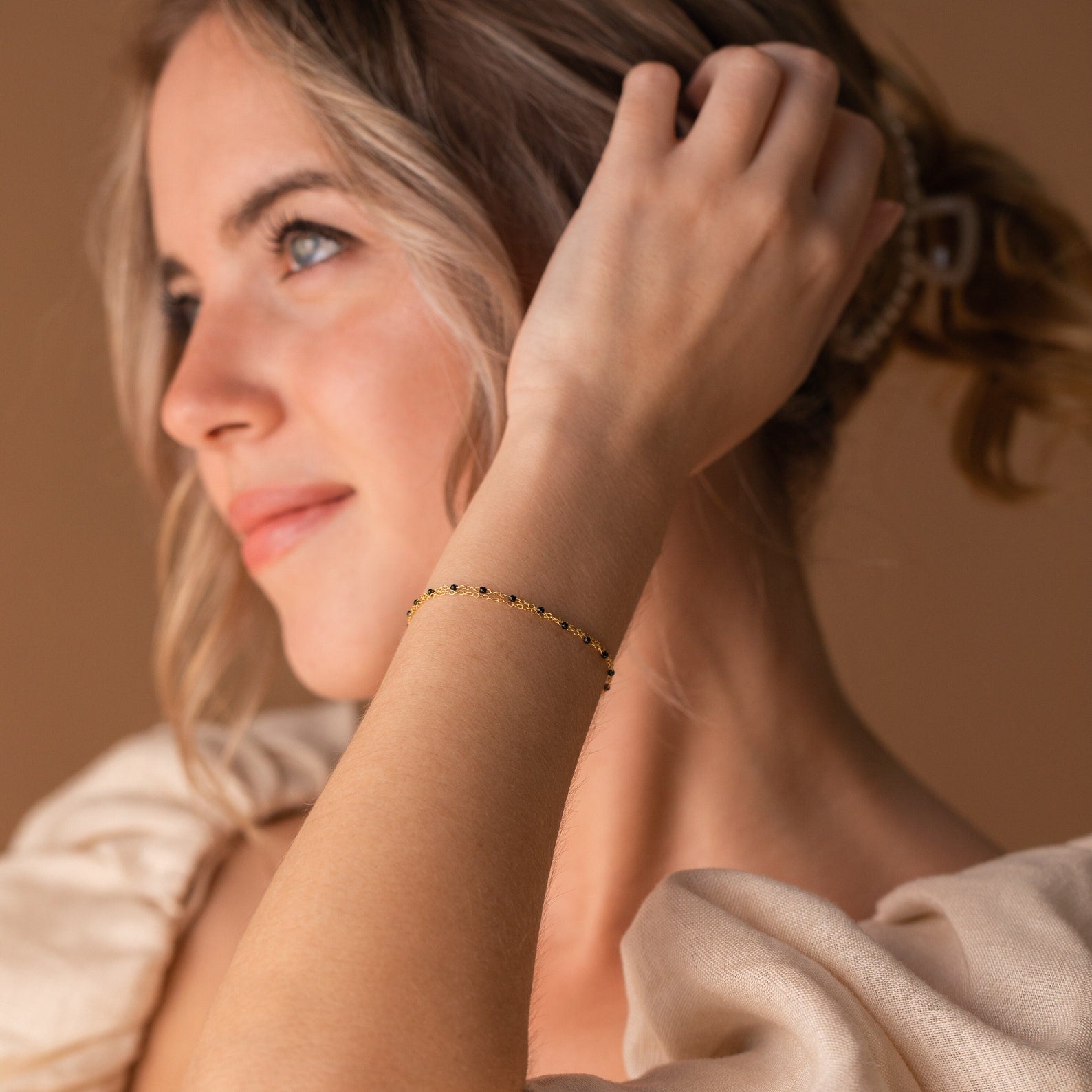 A woman with light hair wears the Calypso Station Bracelet, her hand near her head in soft lighting—an ideal choice for stacking boho accessories.
