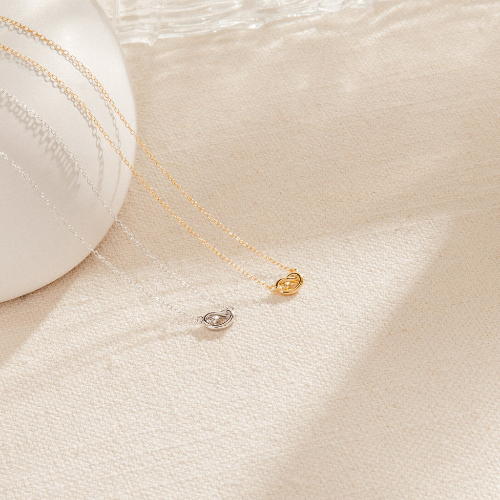 Two delicate Love Knot Necklaces, one in gold and one in silver, are displayed on cream fabric. Each necklace features a small pendant symbolizing unity—making them perfect jewelry gifts for weddings.