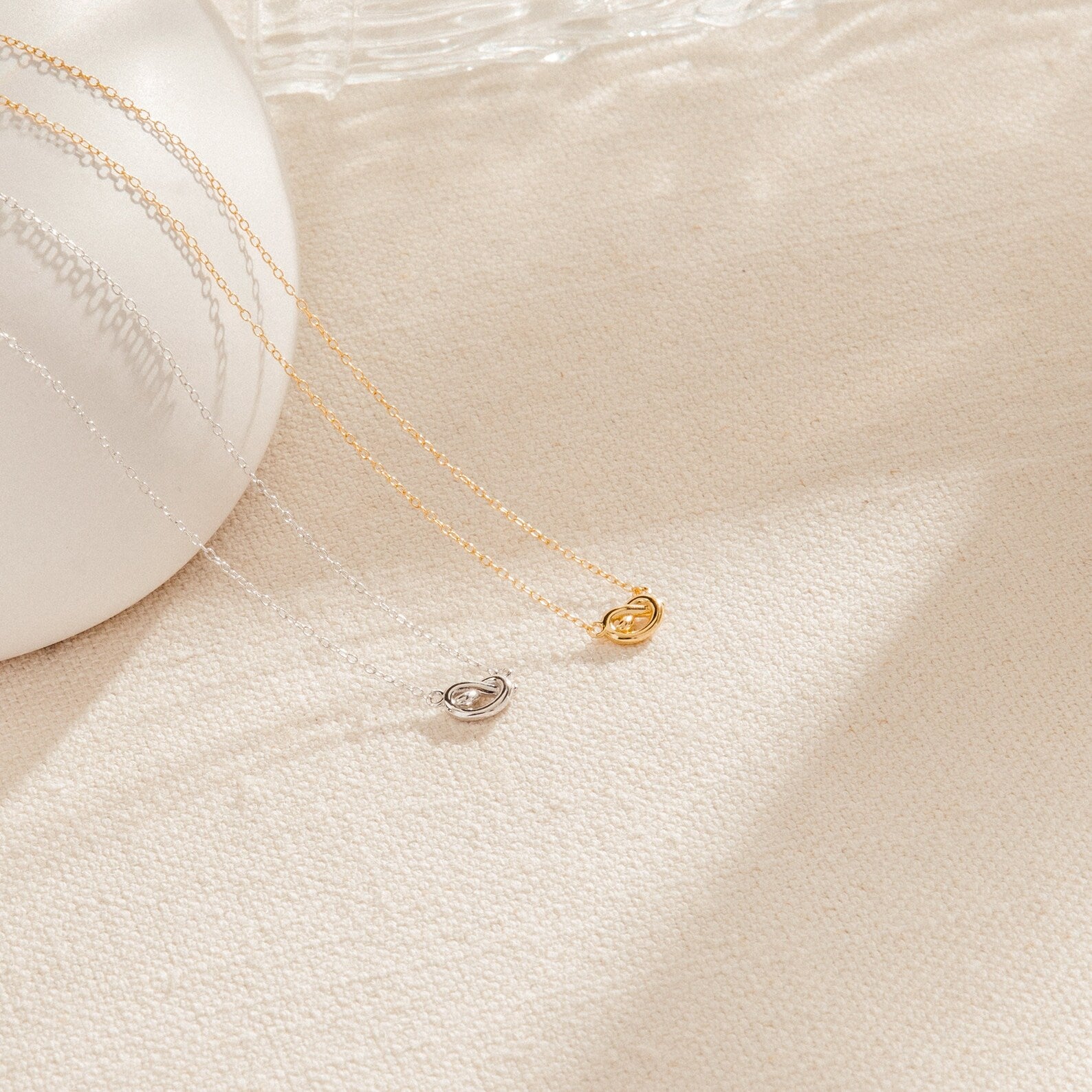 Two delicate Love Knot Necklaces, one in gold and one in silver, are displayed on cream fabric. Each necklace features a small pendant symbolizing unity—making them perfect jewelry gifts for weddings.