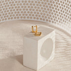 The Kelsey Link Earrings, featuring a contemporary gold square design, rest on a white geometric block for a minimal accessory touch against a softly lit, patterned background.