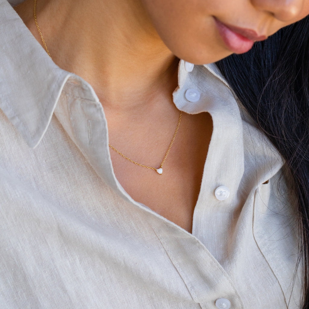 Woman wearing a beige linen shirt and the Madeline Opal Heart Necklace, which features a dainty gold chain and small opal heart-shaped pendant.