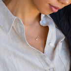 Woman wearing a beige linen shirt and the Madeline Opal Heart Necklace, which features a dainty gold chain and small opal heart-shaped pendant.