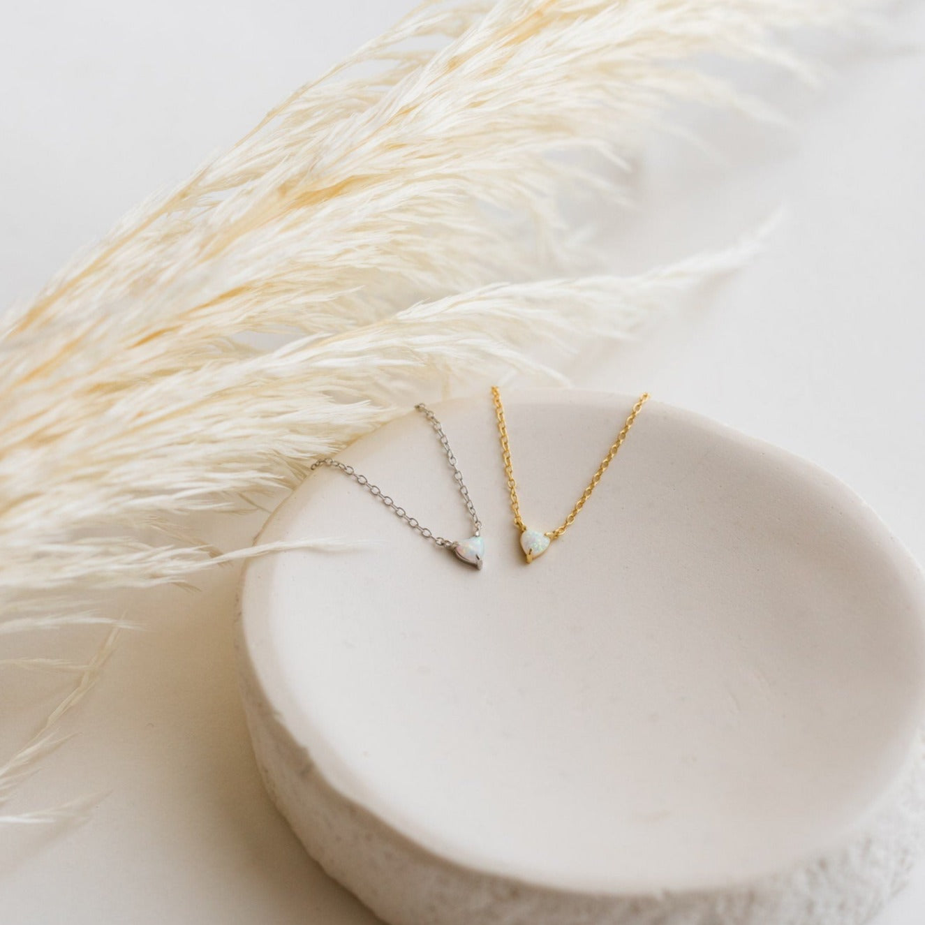 The Madeline Opal Heart Necklace, available in silver and gold, is displayed on a white ceramic dish with pampas grass. Each dainty necklace features a subtle opal heart charm for an elegant touch.