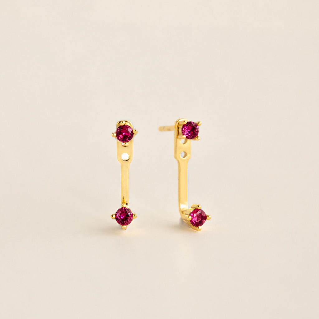 Straight‑on shot of a garnet pair gold birthstone ear jacket set—two small round CZ stones linked by a delicate bar—laid flat on a neutral background.