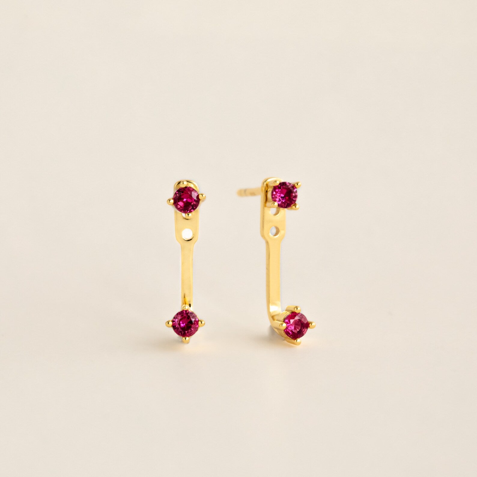 Straight‑on shot of a garnet pair gold birthstone ear jacket set—two small round CZ stones linked by a delicate bar—laid flat on a neutral background.