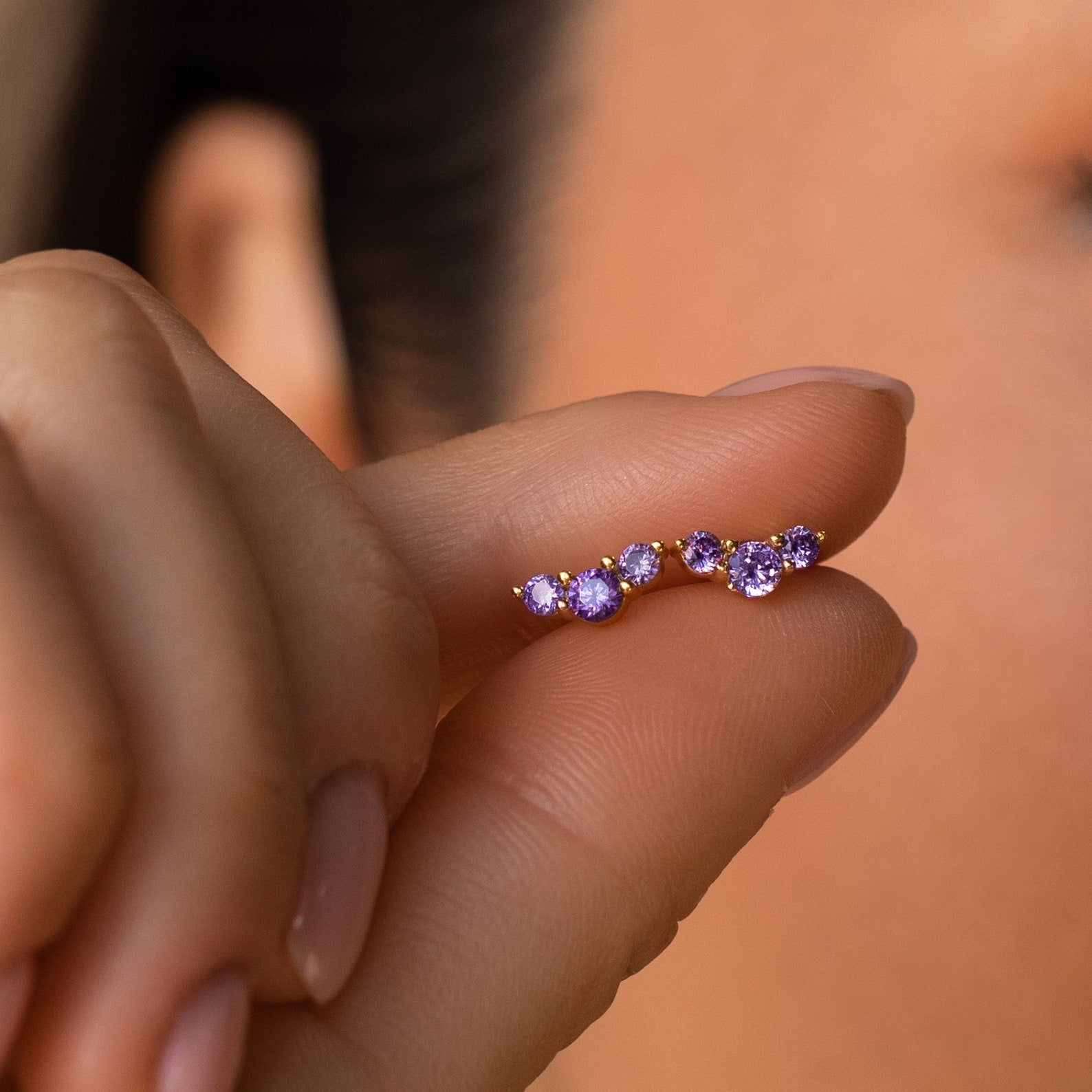 A hand holds a Bella Birthstone Stud earring with five purple gemstones, shown near an ear—an ideal personalized jewelry piece and a meaningful gift for mothers and daughters.