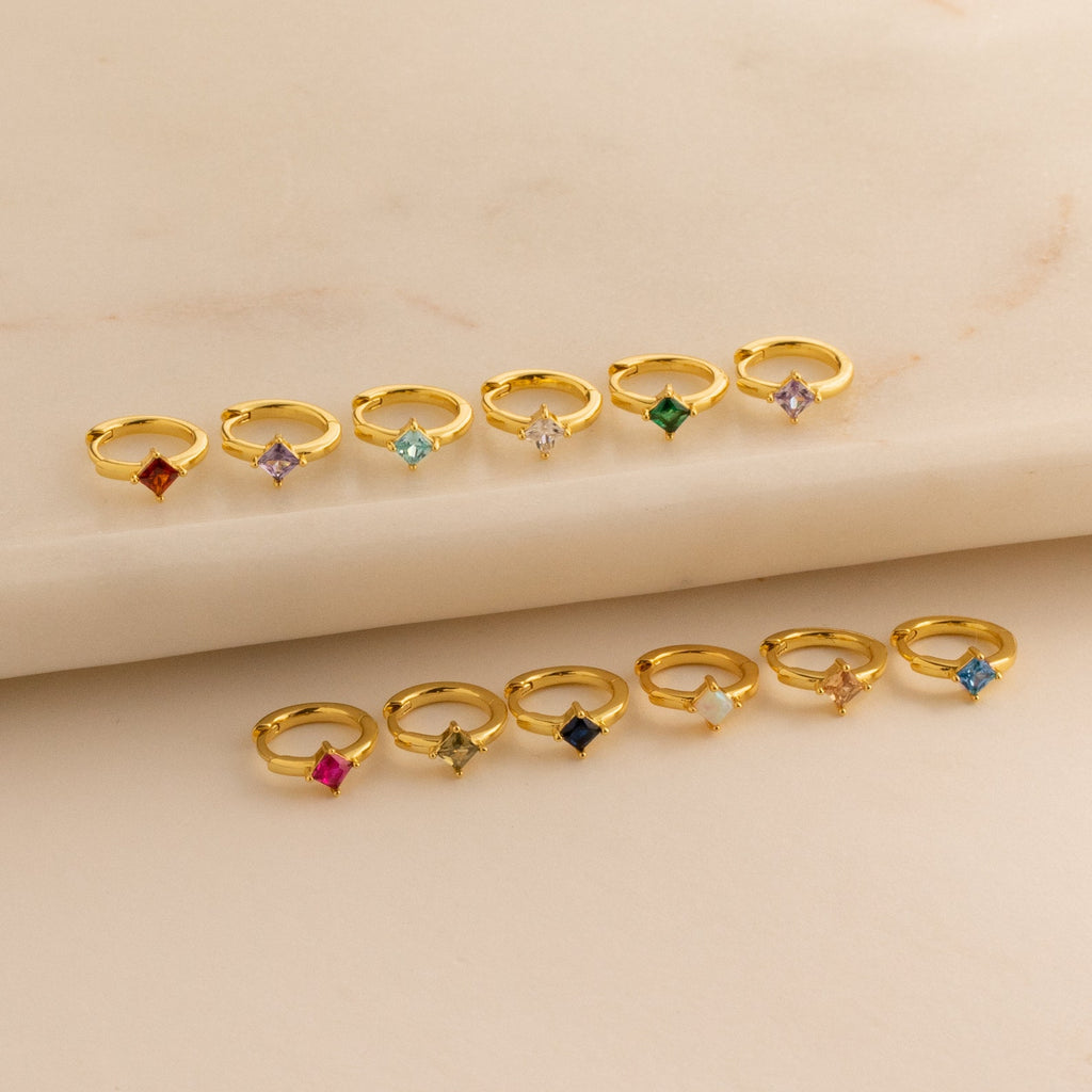 Twelve Athena Opal Huggies are displayed in two rows on a beige marble surface, each gold ring featuring vibrant opal gemstones.