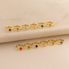 Twelve Athena Opal Huggies are displayed in two rows on a beige marble surface, each gold ring featuring vibrant opal gemstones.