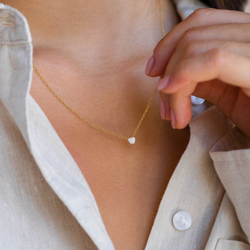 Gold chain necklace with a small heart-shaped white gemstone pendant, worn with a partially unbuttoned beige shirt.