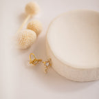 Starry Night Ear Cuffs—gold, star-shaped and adorned with sparkling crystals—rest on a white dish framed by cream dried flowers. These stylish cuff earrings are perfect for anyone seeking a no-piercing option.