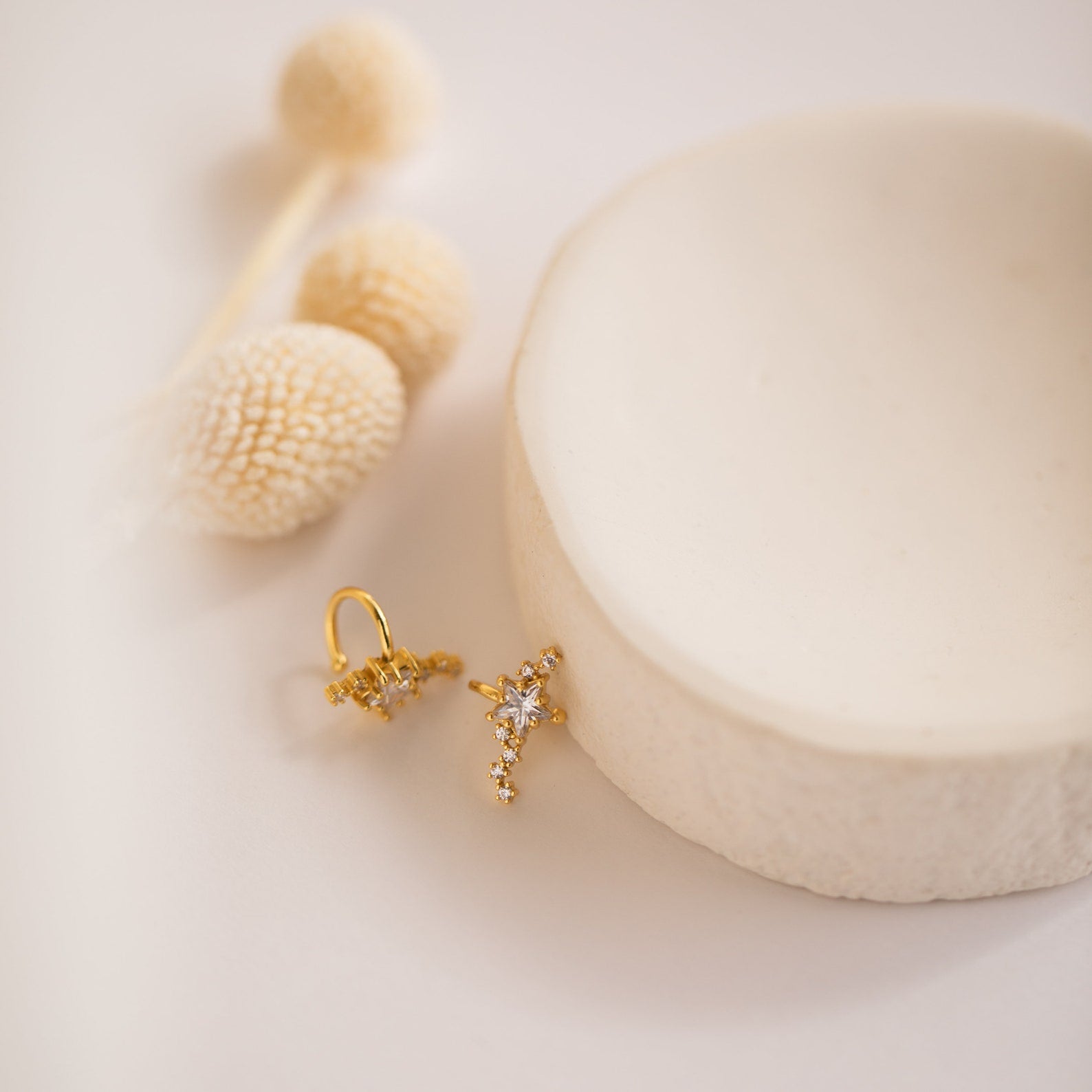 Starry Night Ear Cuffs—gold, star-shaped and adorned with sparkling crystals—rest on a white dish framed by cream dried flowers. These stylish cuff earrings are perfect for anyone seeking a no-piercing option.