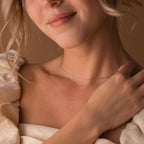 A woman smiles gently, touching her collarbone while wearing the Dainty Chain Necklace and a cream off-shoulder dress, showcasing a minimal style.