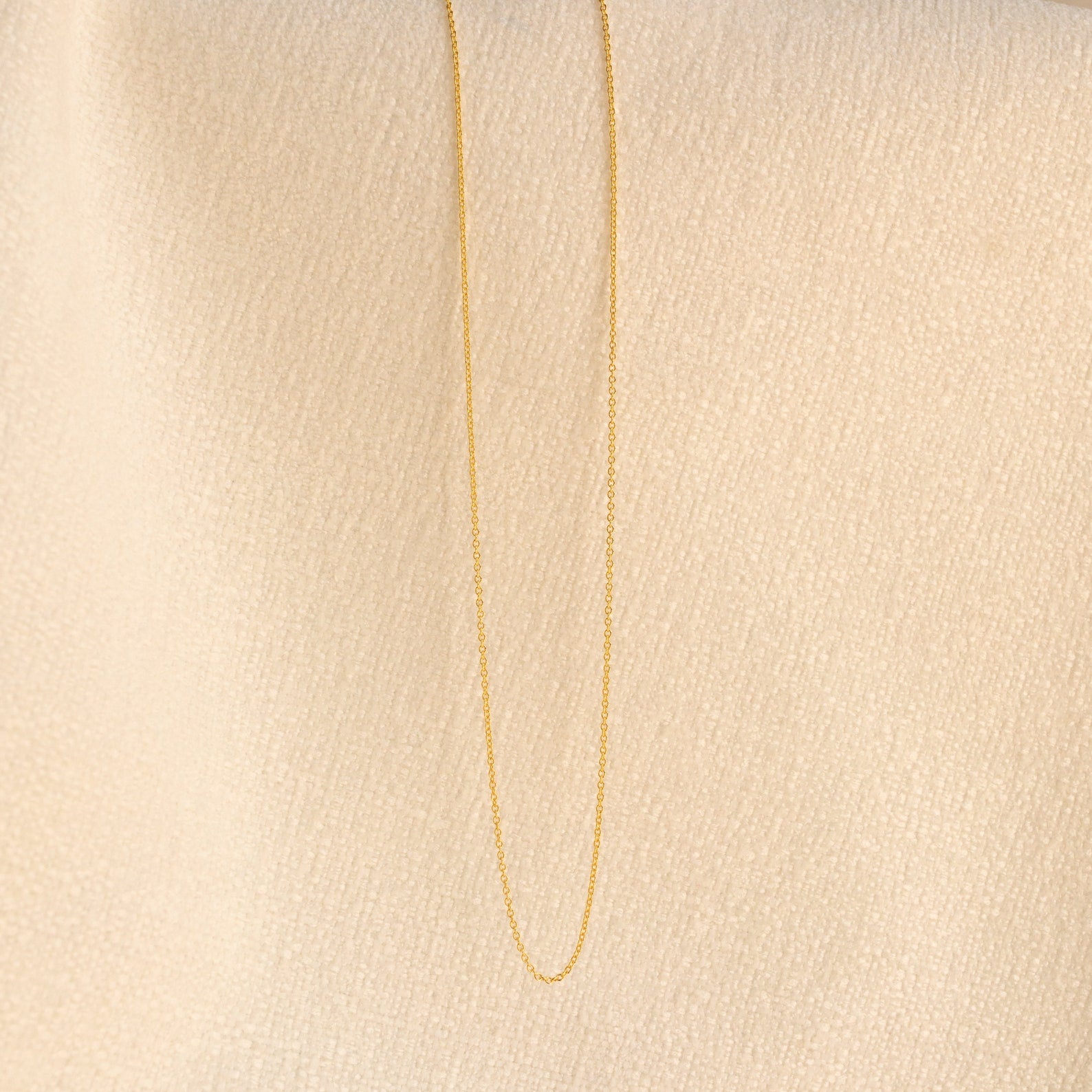 The Dainty Chain Necklace hangs against a textured beige fabric background, ideal for a minimal style or layered with other favorites.