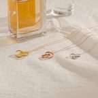 The Engraved Interlocking Necklace features three interlocking circles in gold, rose gold, and silver with personalized engraving, displayed on a white cloth beside a glass perfume bottle—an elegant anniversary gift.