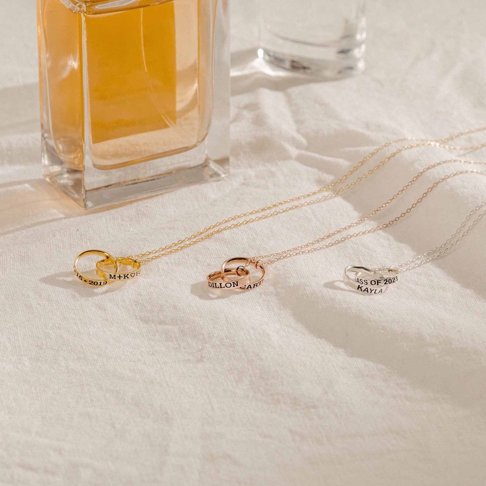 The Engraved Interlocking Necklace features three interlocking circles in gold, rose gold, and silver with personalized engraving, displayed on a white cloth beside a glass perfume bottle—an elegant anniversary gift.