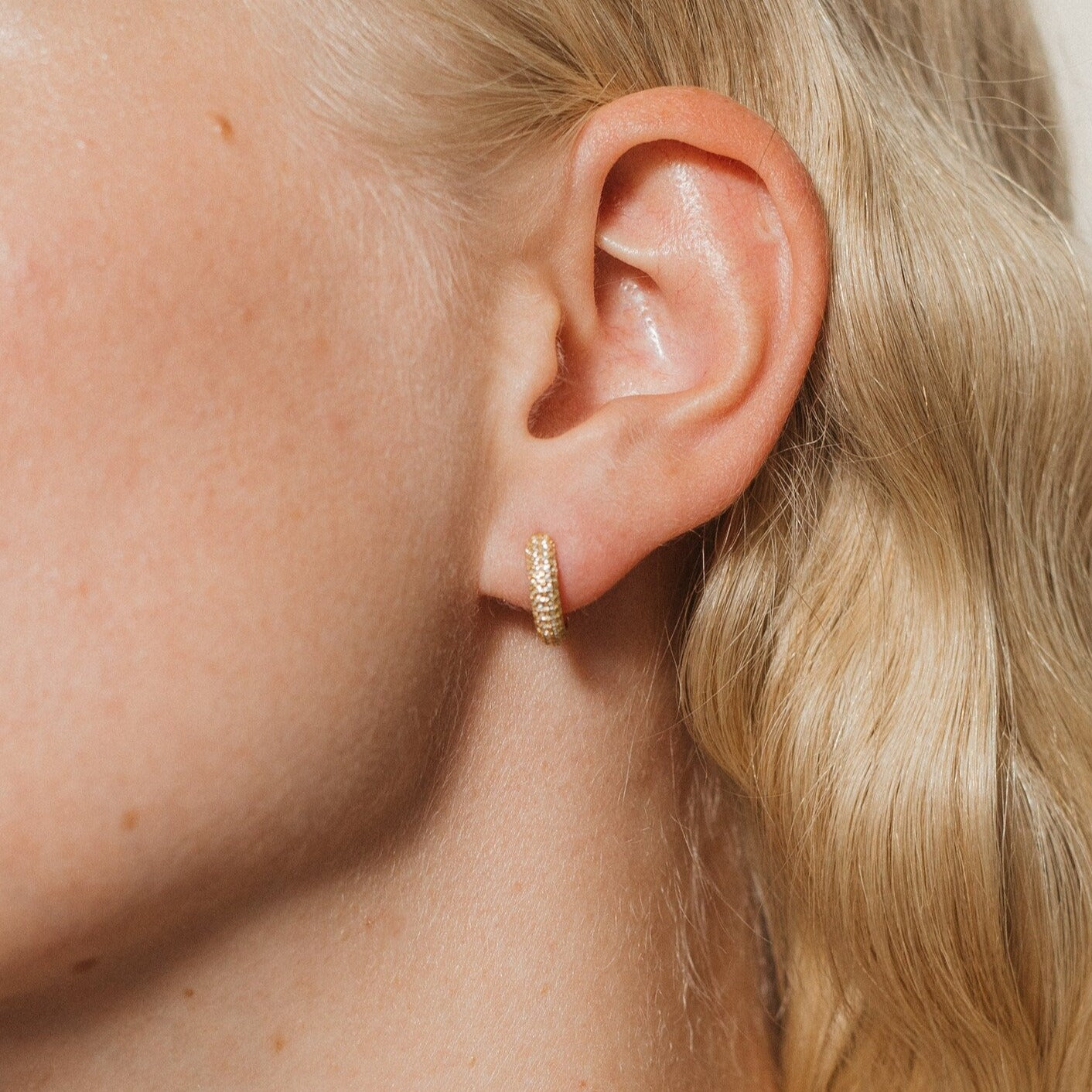 A close-up of a woman's ear shows the Clarissa Pave Huggies—small gold hoops that sparkle against her wavy blonde hair, making them perfect for date night jewelry.