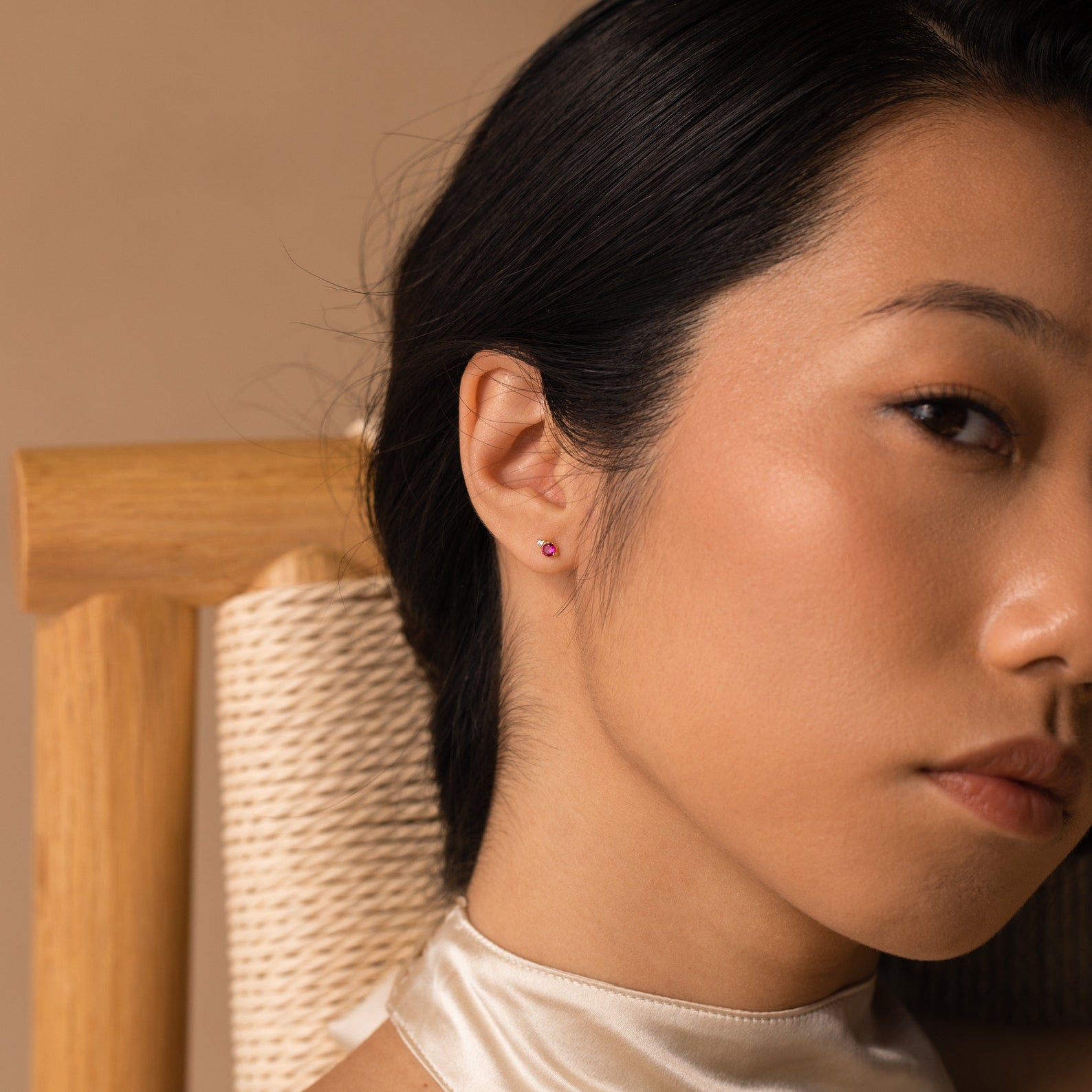 A woman sits in a chair, her look accented by dainty Raindrop Birthstone Studs that add a touch of subtle elegance.
