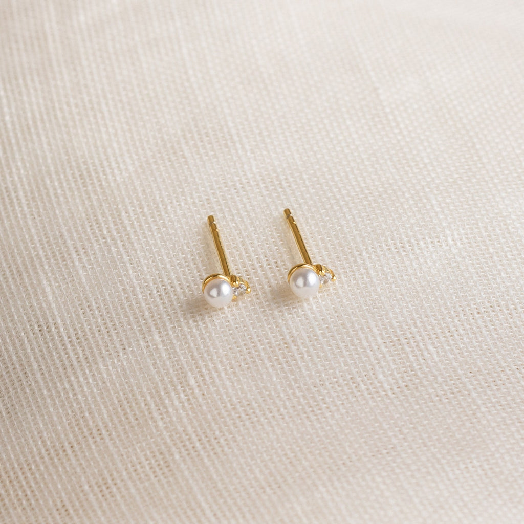 The Raindrop Birthstone Studs, delicate gold earrings adorned with small white pearls, are displayed on a textured cream fabric background.