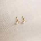 The Raindrop Birthstone Studs, delicate gold earrings adorned with small white pearls, are displayed on a textured cream fabric background.