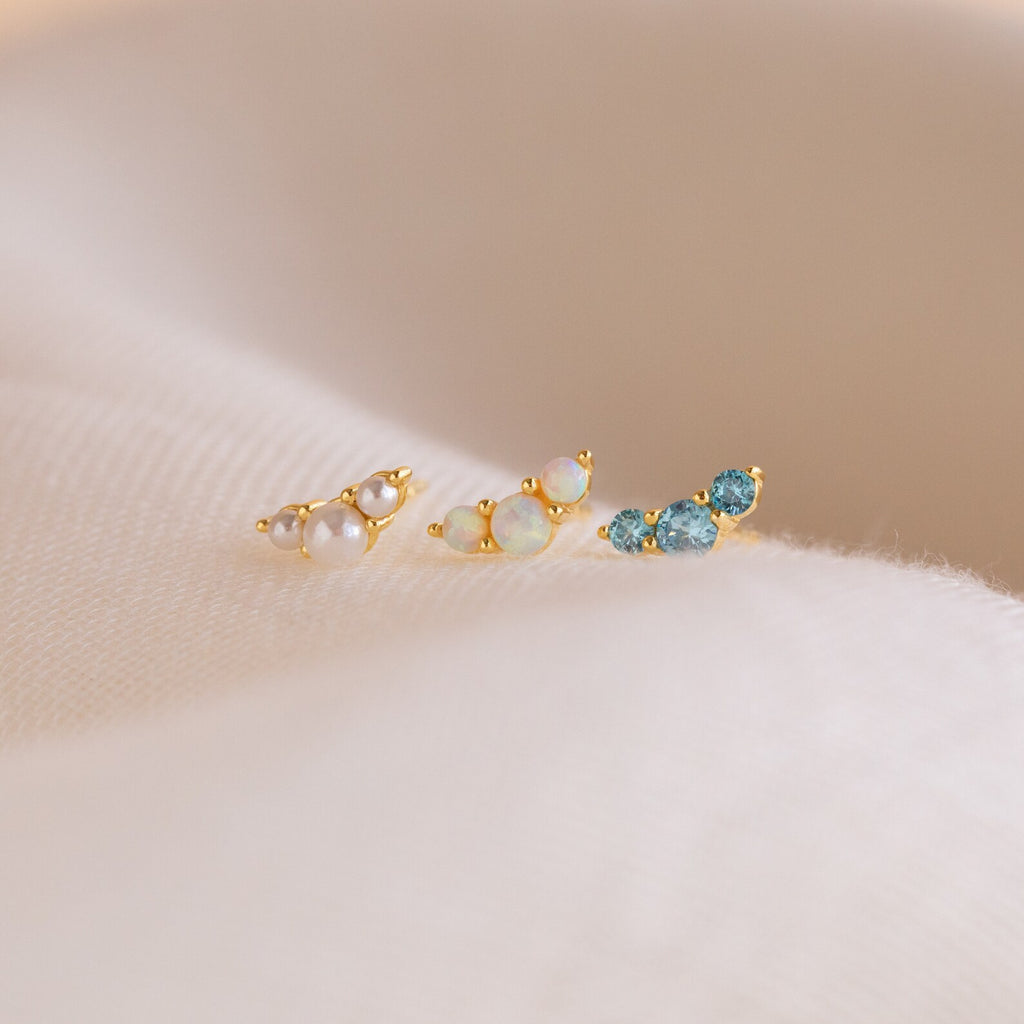 Three Bella Birthstone Studs—delicate gold earrings with pearls, opals, and blue gemstones—make thoughtful gifts for mothers and daughters, displayed on a soft cream fabric background.