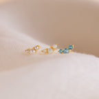 Three Bella Birthstone Studs—delicate gold earrings with pearls, opals, and blue gemstones—make thoughtful gifts for mothers and daughters, displayed on a soft cream fabric background.